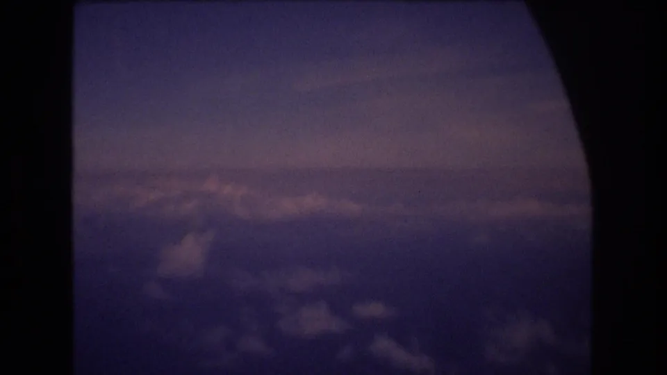 1977 8mm home-movie frame showing clouds and twilight sky seen through a dark airplane window during travel associated with Honolulu, Hawaii metadata.