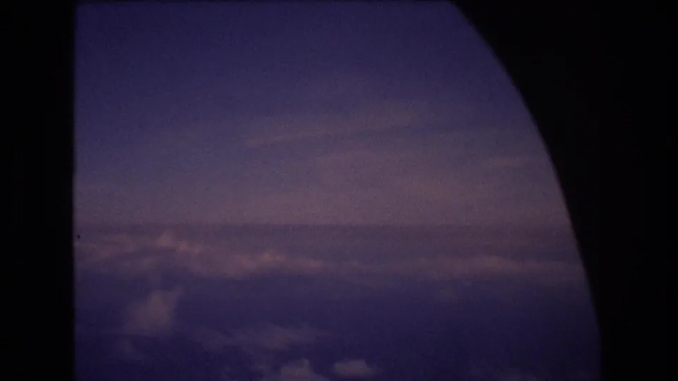 A purple-blue aerial view over clouds appears through the curved airplane window frame in this 1970s 8mm travel shot.