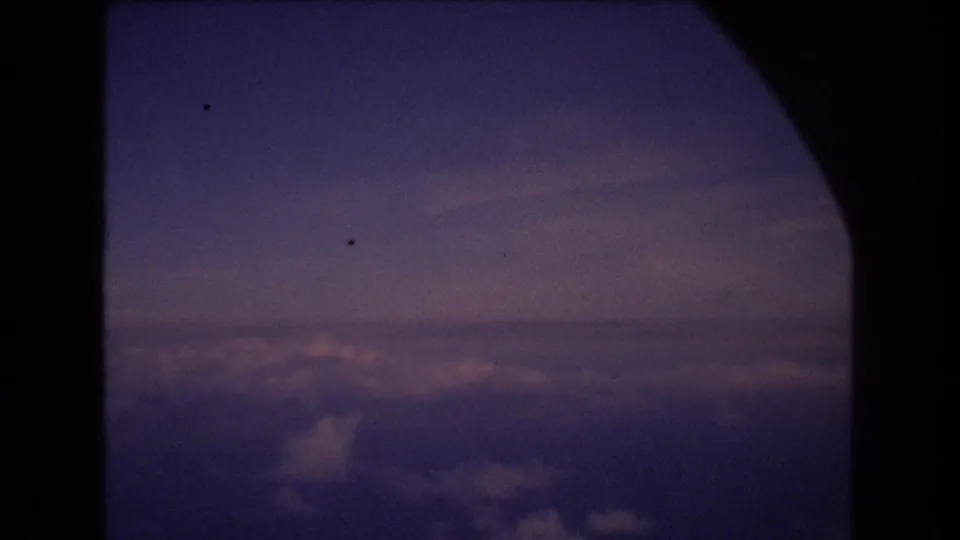The frame shows a passenger-window view of clouds below a muted twilight sky, with the aircraft interior reduced to dark silhouettes.
