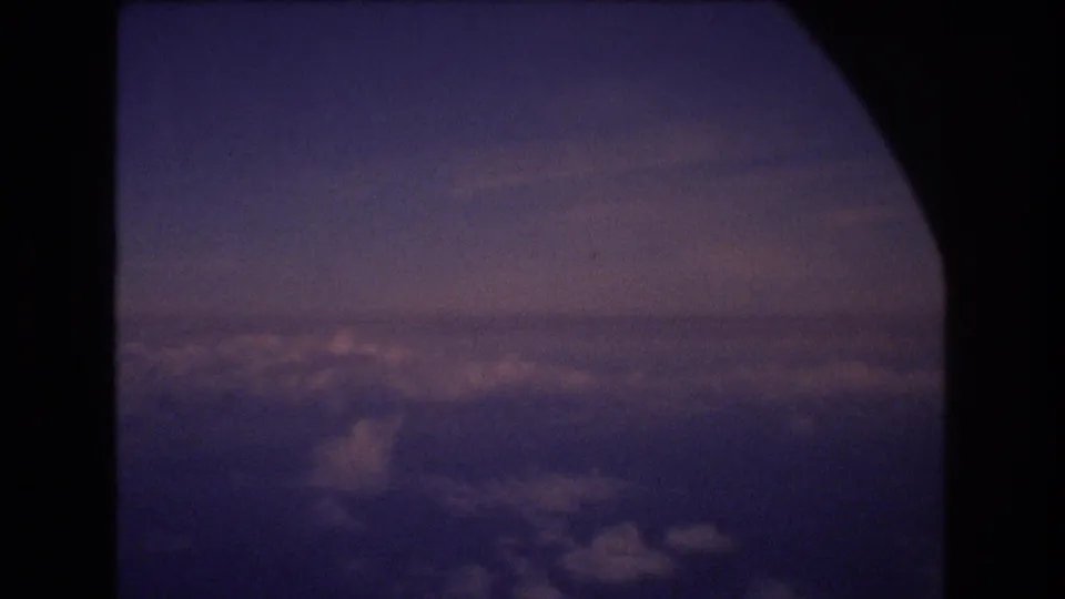 Clouds and a distant horizon sit beneath a purple-blue sky, framed by the dark curve of an airplane window in 8mm home-movie footage.