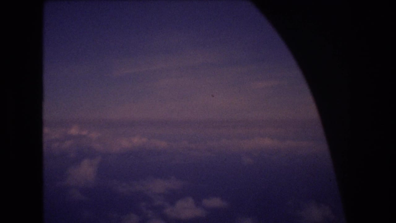 1977 8mm home-movie frame from an airplane window, showing a dark rounded window edge and purple-blue clouds below a dim horizon.