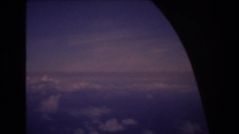 1977 8mm home-movie frame from an airplane window, showing a dark rounded window edge and purple-blue clouds below a dim horizon.