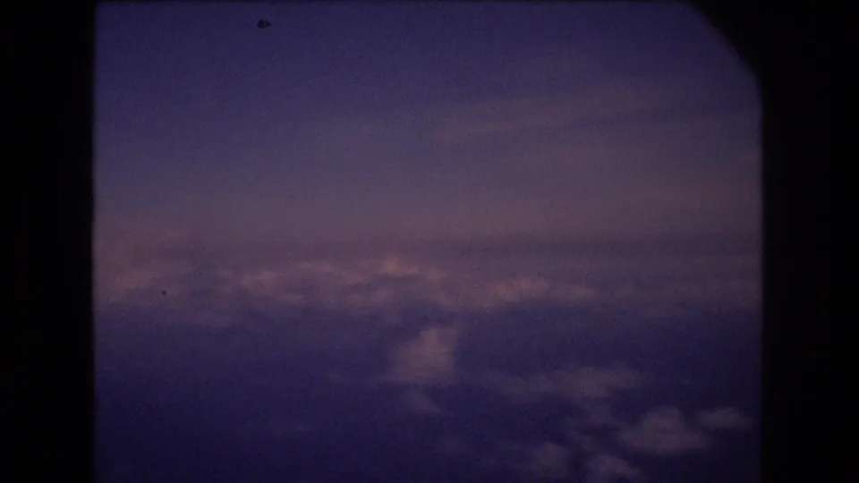 In-flight view through a dark aircraft window frame, with layered clouds and subdued purple twilight tones supported by the 1977 Honolulu travel metadata.