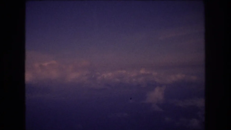 8mm passenger-window view with a dark vertical frame edge, cloud tops below, and a muted blue-purple sky in a brief 1977 travel clip.