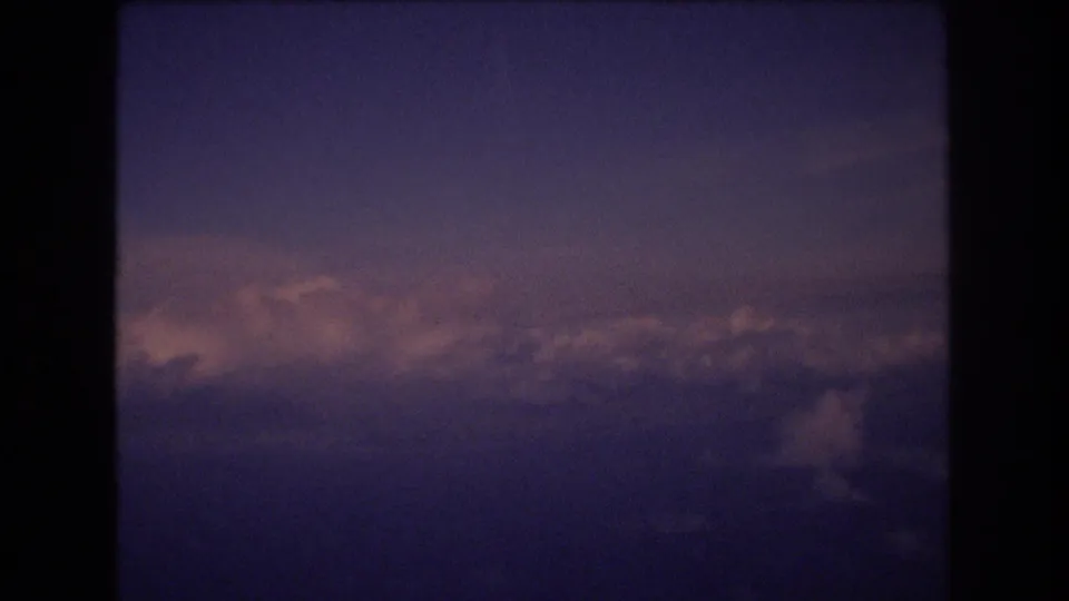 Final frame of the 1977 8mm in-flight view, with dark aircraft-window framing around a softly lit cloudscape and purple-blue sky.