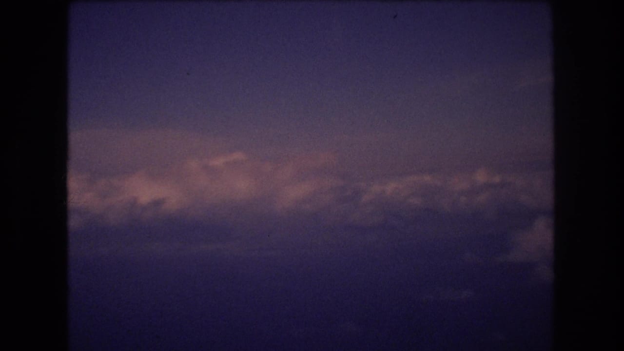 1977 8mm travel footage frame showing a purple-blue dusk sky above a horizontal bank of softly lit clouds during an airplane-window view associated with Honolulu travel metadata.