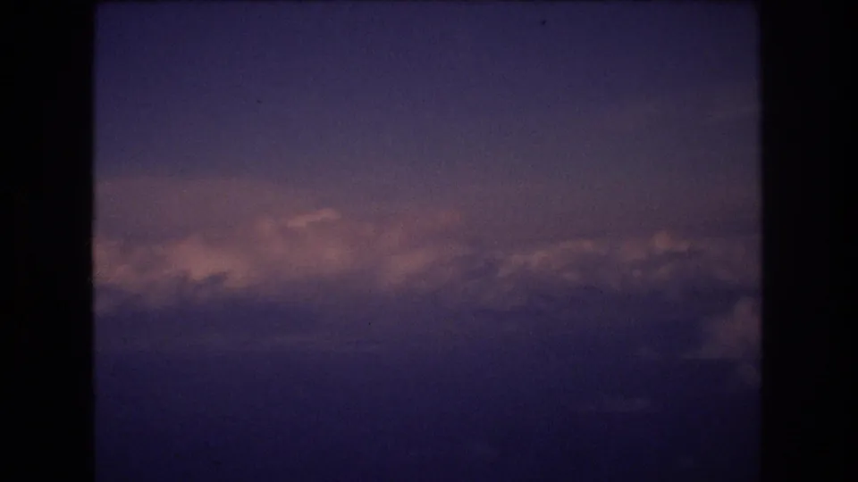 1977 8mm travel footage frame showing a purple-blue dusk sky above a horizontal bank of softly lit clouds during an airplane-window view associated with Honolulu travel metadata.