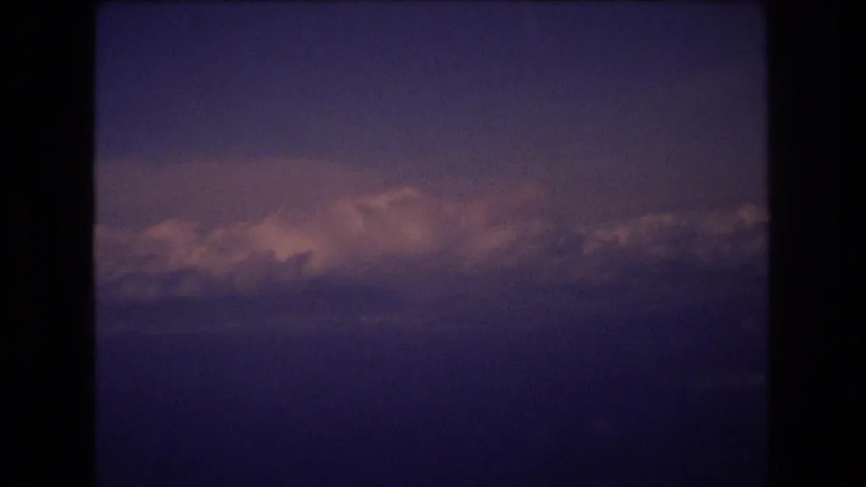 A 1977 8mm frame shows layered clouds under a deepening purple sky, with pink-orange sunset highlights and the rounded edge of the scanned home-movie frame.