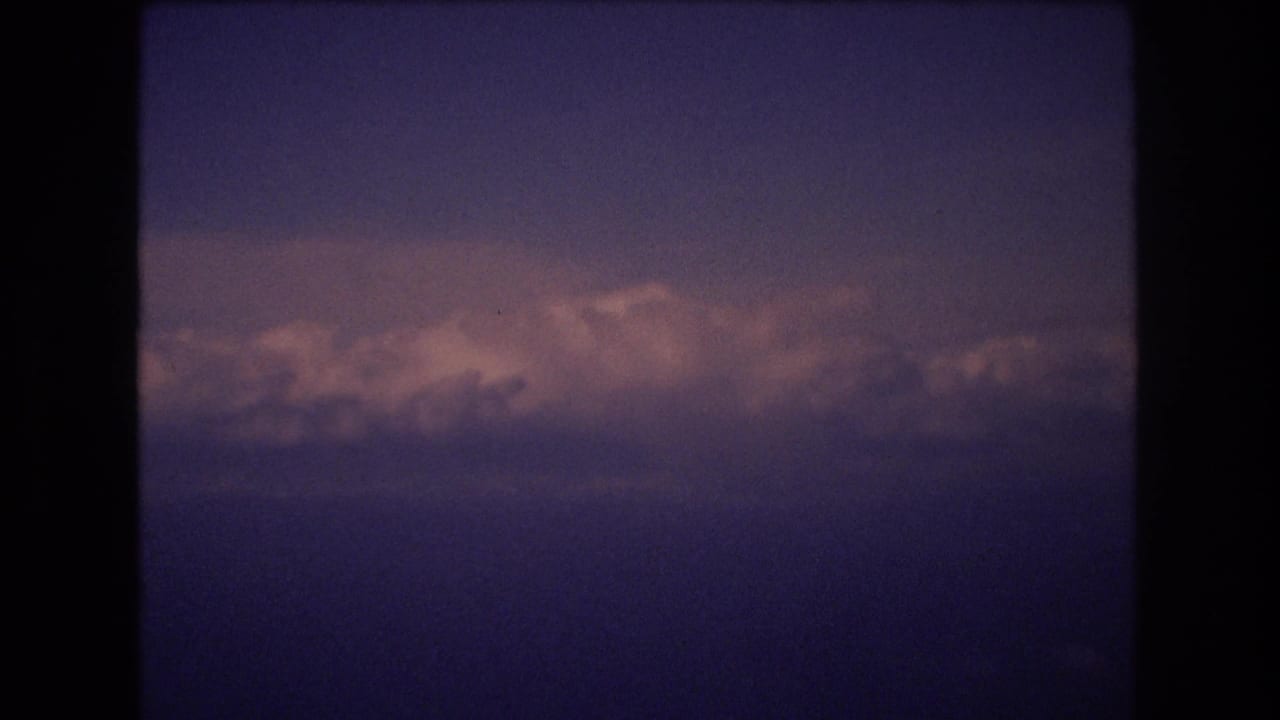 1977 8mm home-movie frame associated with Honolulu travel footage, showing layered clouds lit by pinkish light against a purple-blue sky with dark rounded borders.