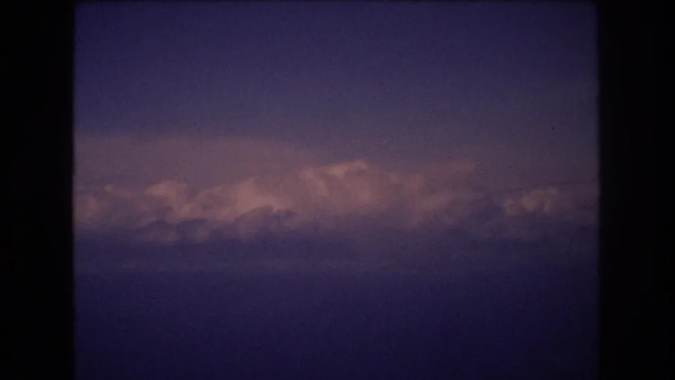 A 1977 8mm travel frame shows a broad cloud bank and muted pink illumination across a dark blue-purple sky, with the film edge visible around the image.