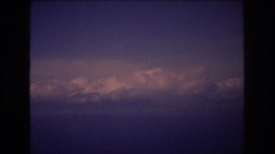 Grainy 8mm home-movie frame from the Honolulu-associated clip shows a low band of clouds beneath a darker blue-purple sky, with strong dark border areas from the film scan.