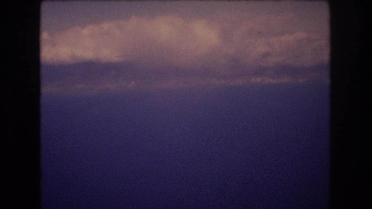 1977 Honolulu metadata-supported 8mm travel frame showing a blue ocean or lake horizon through dark aircraft-window framing with clouds above.