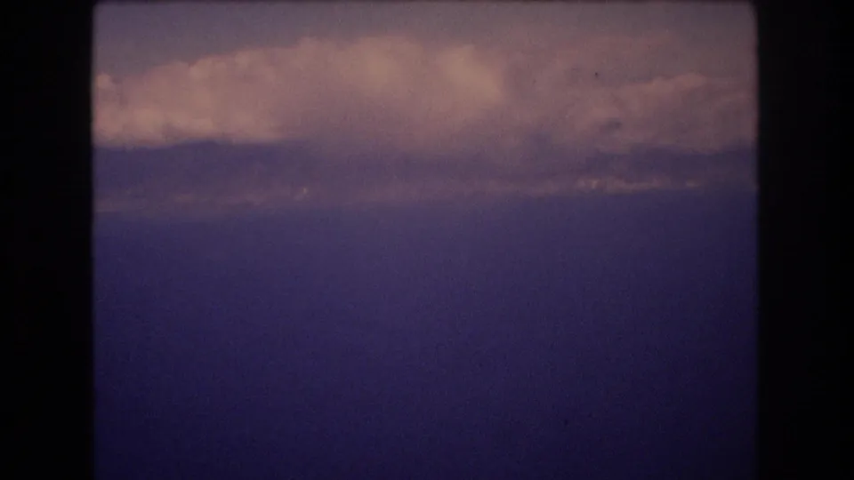 1977 Honolulu metadata-supported 8mm travel frame showing a blue ocean or lake horizon through dark aircraft-window framing with clouds above.