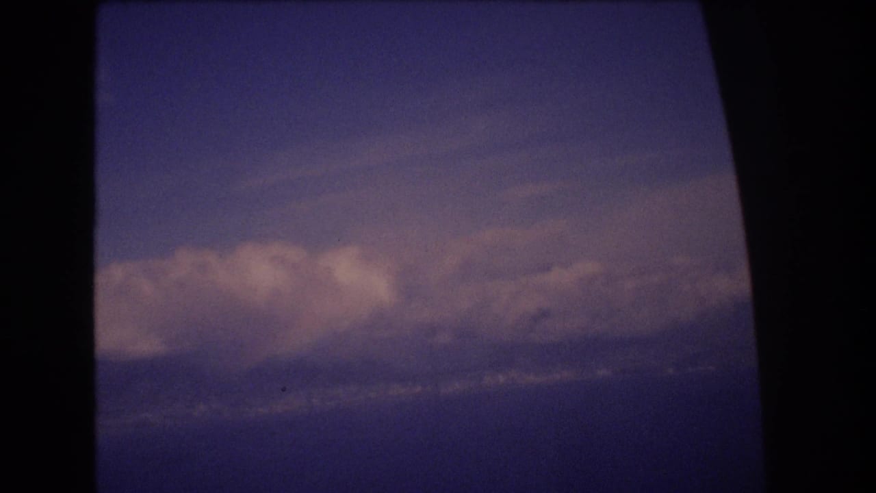 1977 8mm travel footage shows blue sky, distant white clouds, and a dark rounded window edge during a flight associated with Honolulu, Hawaii.
