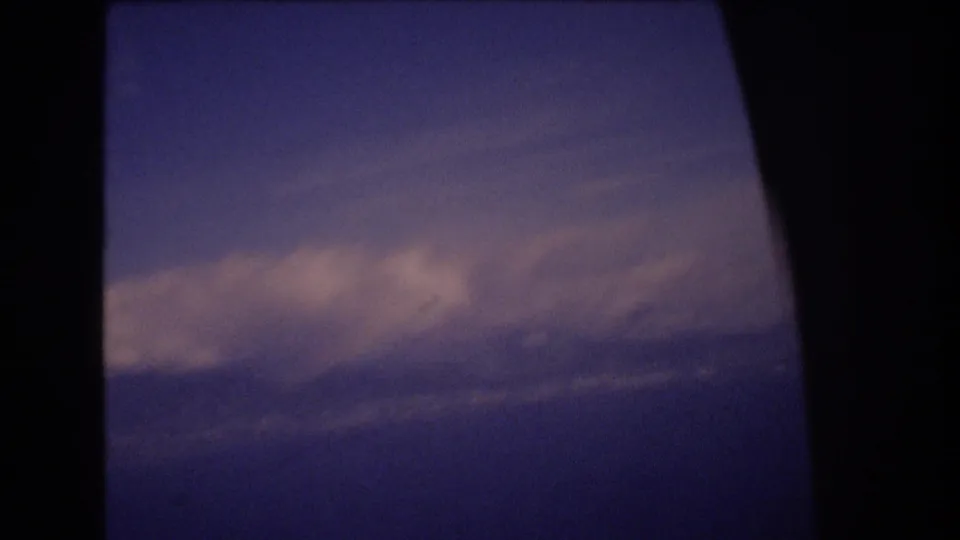 A 1977 8mm frame from the Honolulu travel clip shows layered clouds and blue sky partially bordered by dark airplane-window or frame edges.