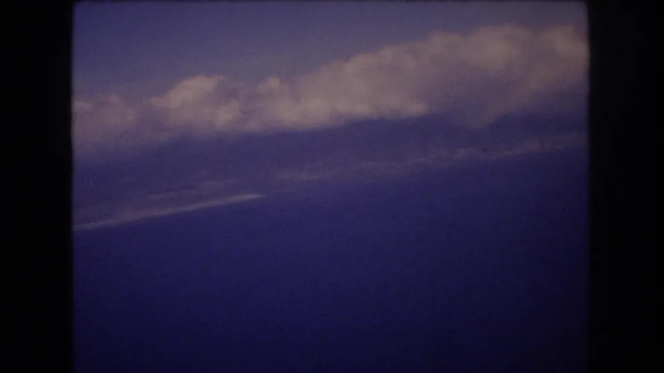 The home-movie frame shows a pale cloud bank over a deep blue aerial view, with dark rounded framing from the plane-window or film contact sheet edge.