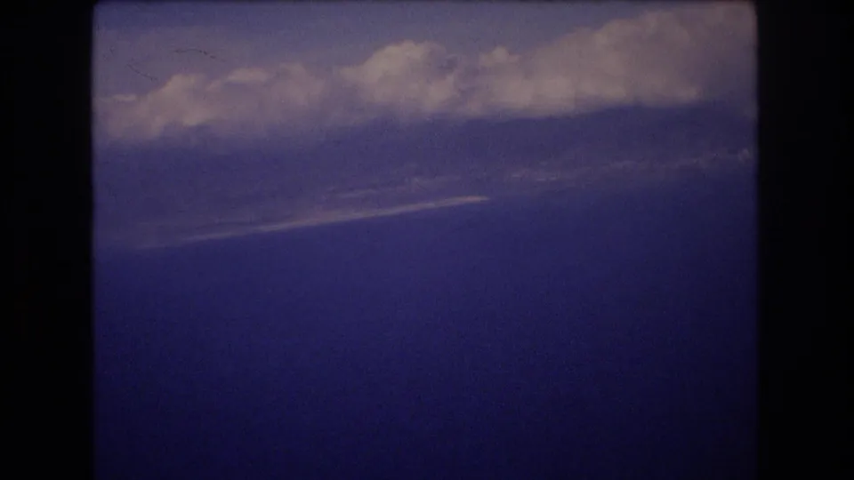 The final 1977 8mm still holds on white clouds and a blue aerial horizon, framed by dark vertical edges from the airplane-window view or projected-film border.