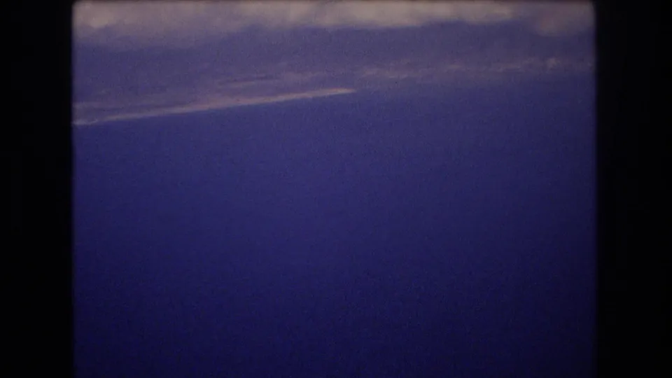 1977 Honolulu travel-footage frame with thick pale clouds across the upper portion and saturated blue sky filling the lower frame.