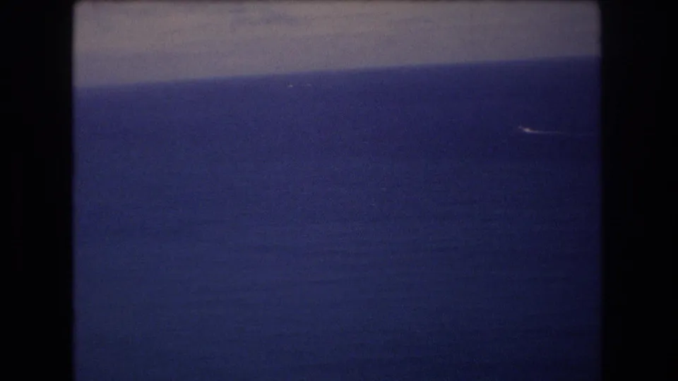 1977 8mm frame from a high vantage near Honolulu showing blue Pacific water, a pale horizontal band, and a tiny white streak that may be a distant boat, aircraft, or wake.