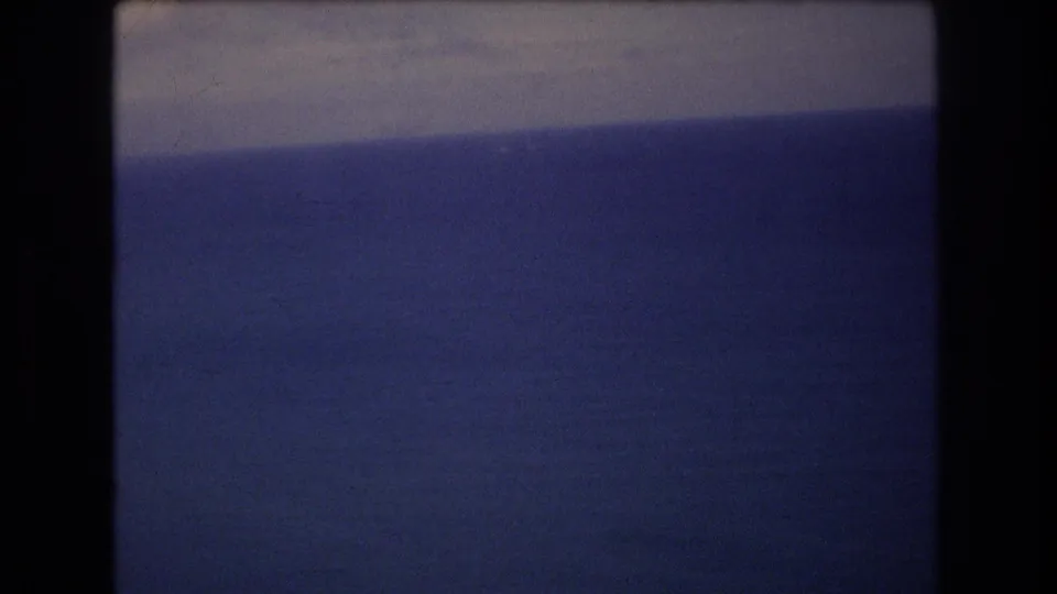 1977 8mm home-movie frame showing deep blue ocean and a pale sky during an aerial approach near Honolulu, with dark vertical frame edges visible.