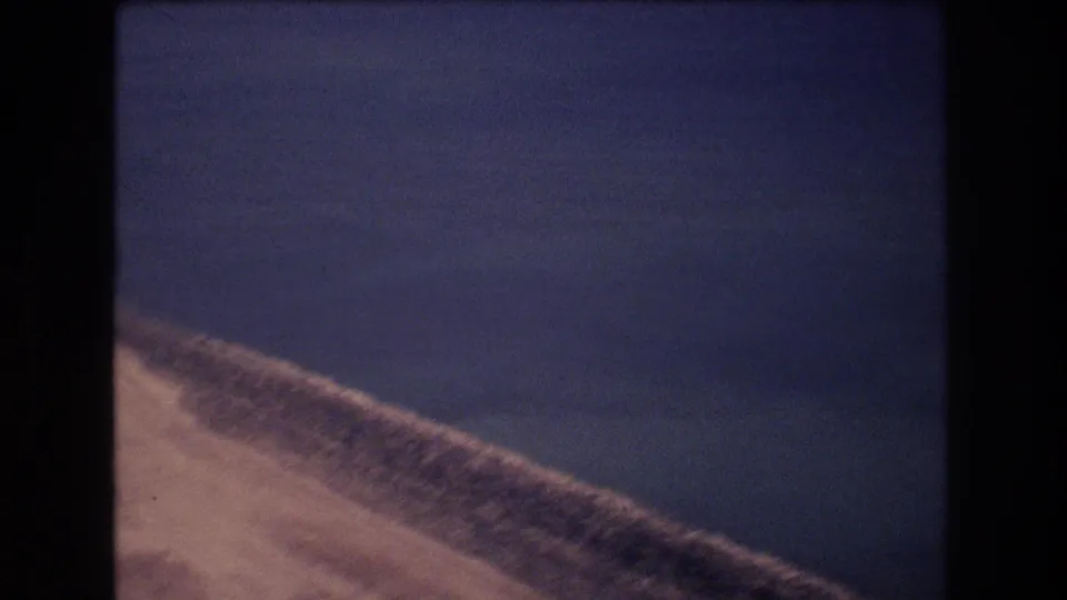 1977 8mm aerial frame near Honolulu showing a sandy shoreline entering the lower frame beneath a broad expanse of blue ocean.
