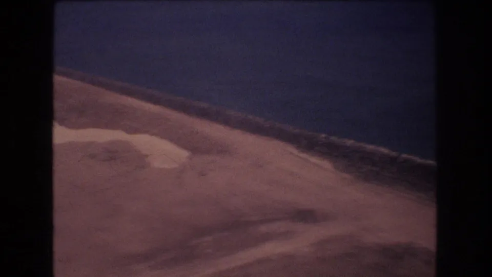 1977 Honolulu-area aerial descent frame showing sandy land, a narrow surf line, and open ocean through the soft, color-faded look of 8mm film.