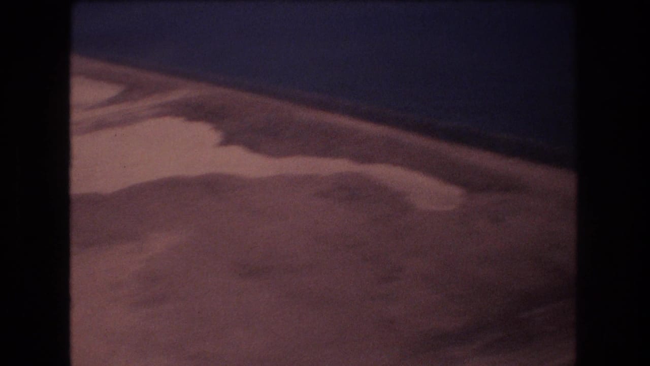 1977 Honolulu-area 8mm frame looking down at pale wet sand, shallow pools, and a dark blue water edge from an aerial perspective.