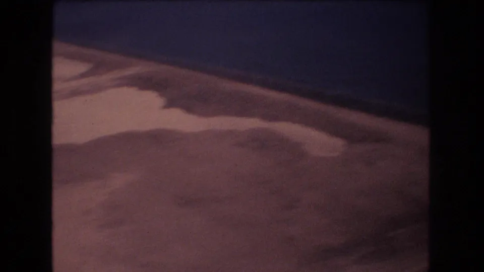 1977 Honolulu-area 8mm frame looking down at pale wet sand, shallow pools, and a dark blue water edge from an aerial perspective.