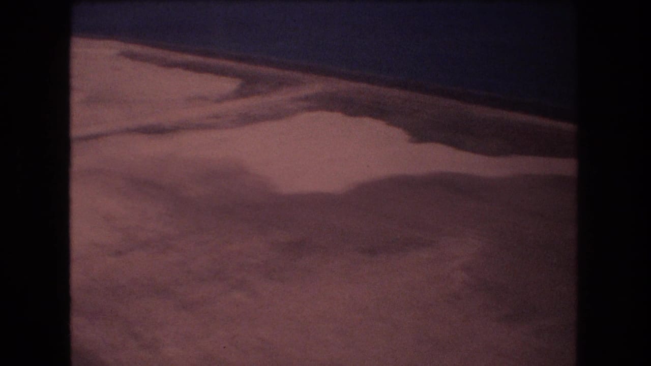 1977 Honolulu-area 8mm aerial frame showing a broad pale shoreline, shallow water shapes, and a dark ocean edge with warm film color.