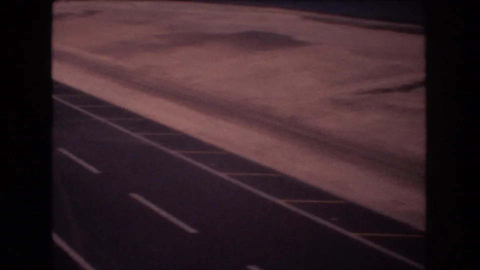 1977 Honolulu-area 8mm aerial frame showing coastal sand and shallow water beside a paved roadway or runway-like surface with white lane markings and yellow diagonal striping.