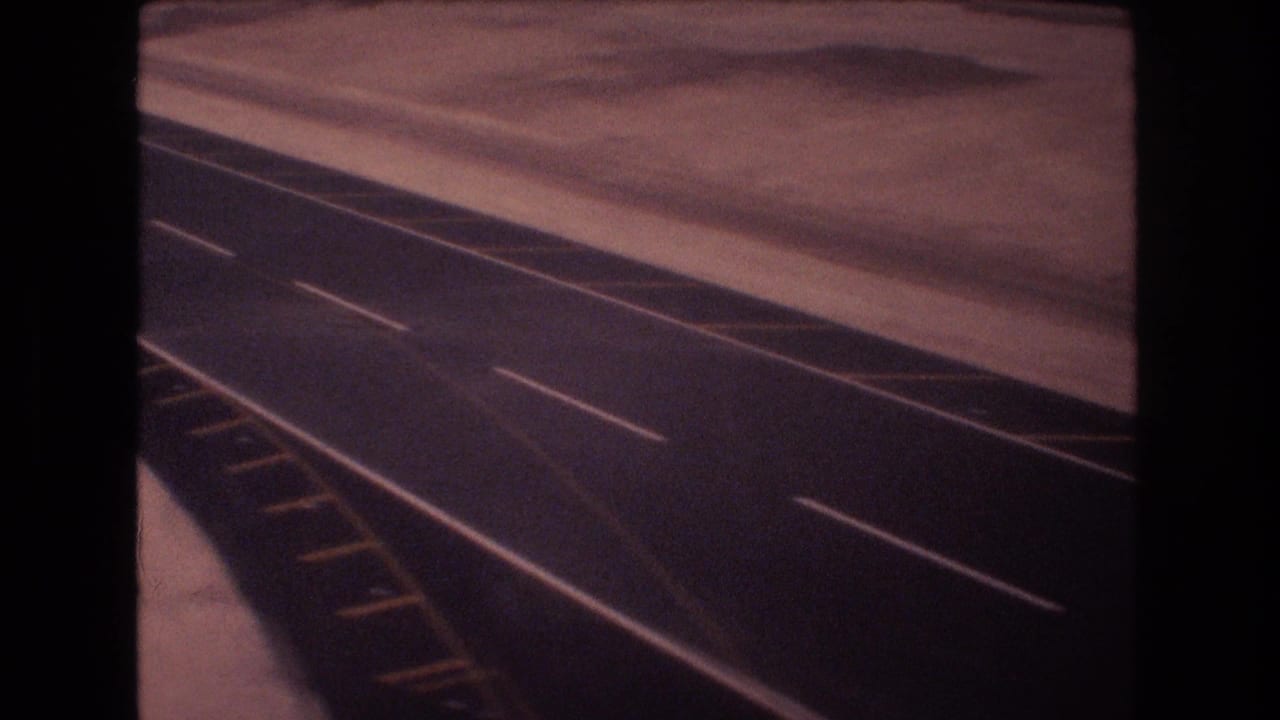 1977 8mm home-movie frame showing a long dark road or runway surface with white dashed lane markings and yellow edge markings in a flat, arid-looking area.