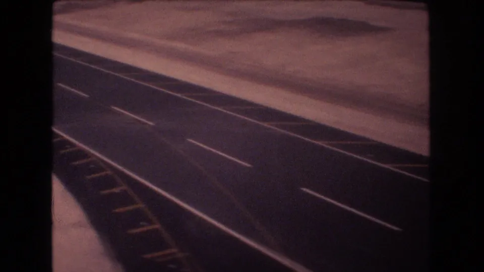 1977 8mm home-movie frame showing a long dark road or runway surface with white dashed lane markings and yellow edge markings in a flat, arid-looking area.