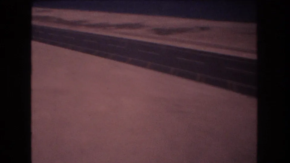Soft 8mm frame showing the dark paved surface continuing through an open flat area, with dashed white markings blurred by motion and film grain.
