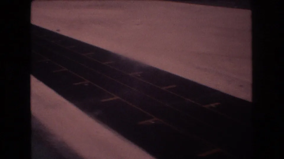 Final 1977 8mm frame showing an empty asphalt road or runway bordered by pale open ground, with yellow markings and a hazy, low-contrast horizon.