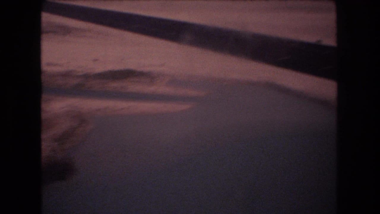 A soft, grainy 8mm frame shows pale open ground, faint uneven terrain, and a dark diagonal edge or shadow as the camera moves through the landscape.