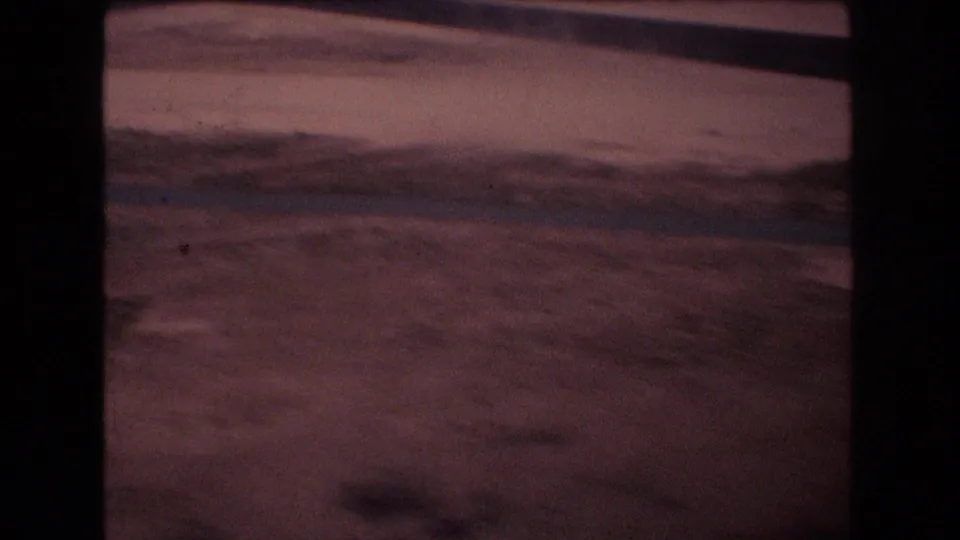 Grainy 1977 home-movie frame with a low view across dusty ground, a dark road or vehicle-edge line, and warm magenta film color.