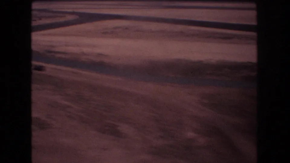 A low moving 8mm view of open, dry-looking terrain with dark linear tracks or shadows and a hazy horizon from the supplied Honolulu Hawaii 1977 reel.