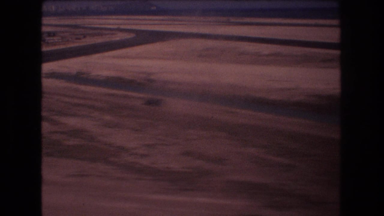 1977 Honolulu airport approach footage showing runway pavement and dusty open ground through a dark, window-like frame on faded 8mm film.