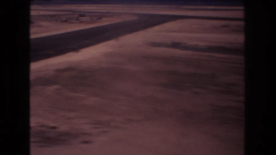 Aircraft-window view over runway pavement and barren airport terrain during a 1977 Honolulu landing approach, with dark film or window borders visible.