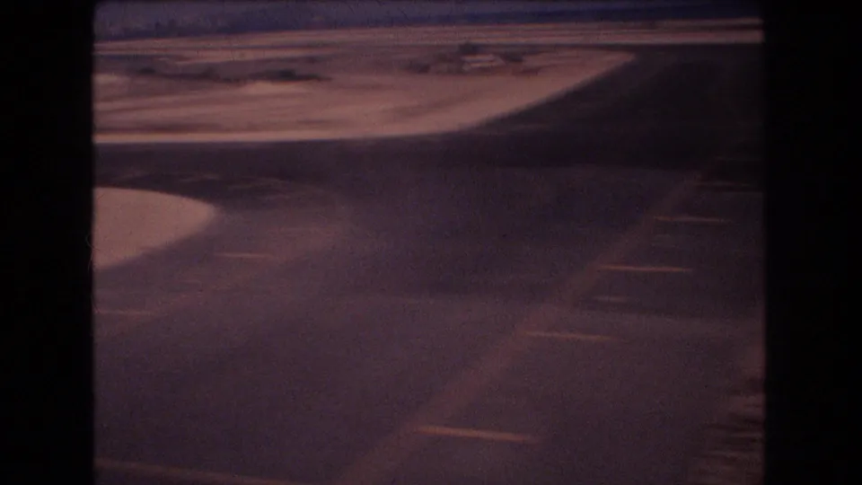 A wing or aircraft structure crosses the frame as the plane descends over runway pavement and dry Honolulu airport ground on grainy 8mm footage.