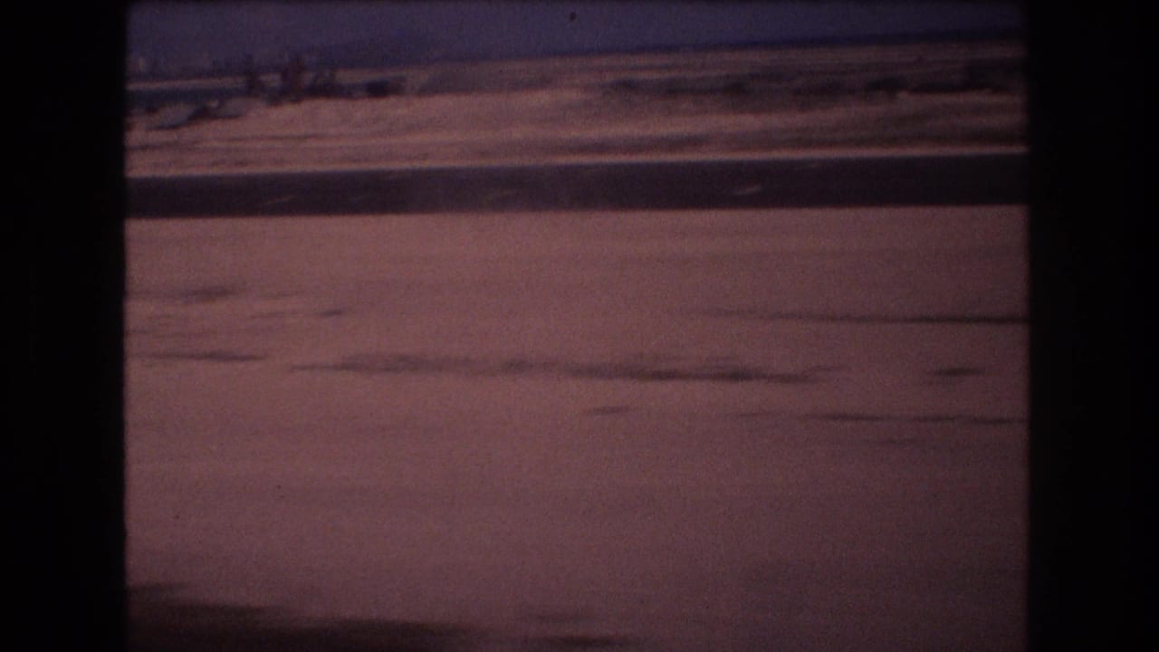 1977 8mm Honolulu-area travel footage appears to show a flat dusty roadside seen from a moving vehicle, with distant low structures and a pink-purple sky.