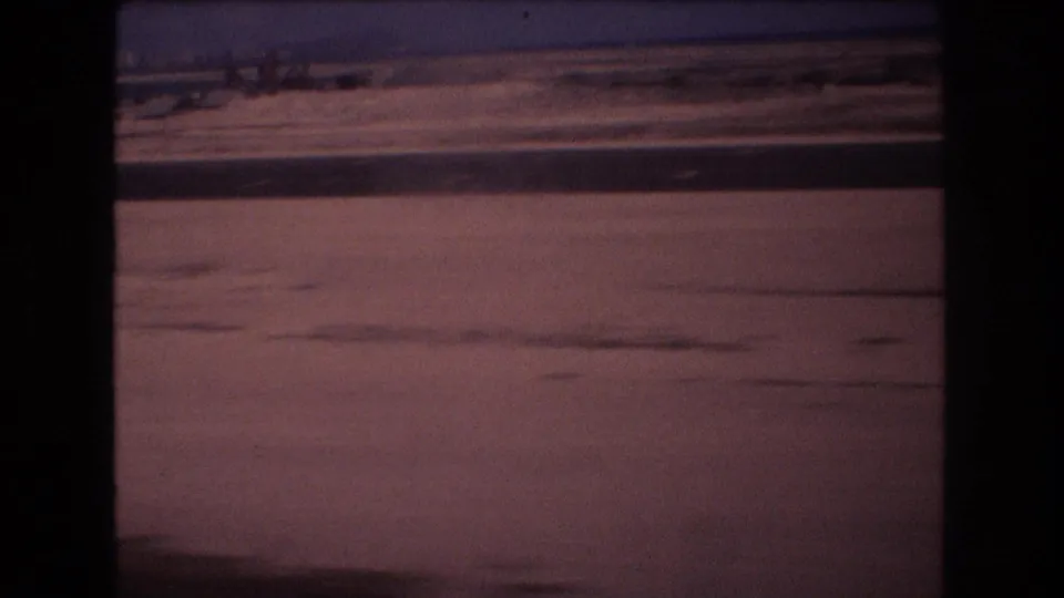 1977 8mm Honolulu-area travel footage appears to show a flat dusty roadside seen from a moving vehicle, with distant low structures and a pink-purple sky.