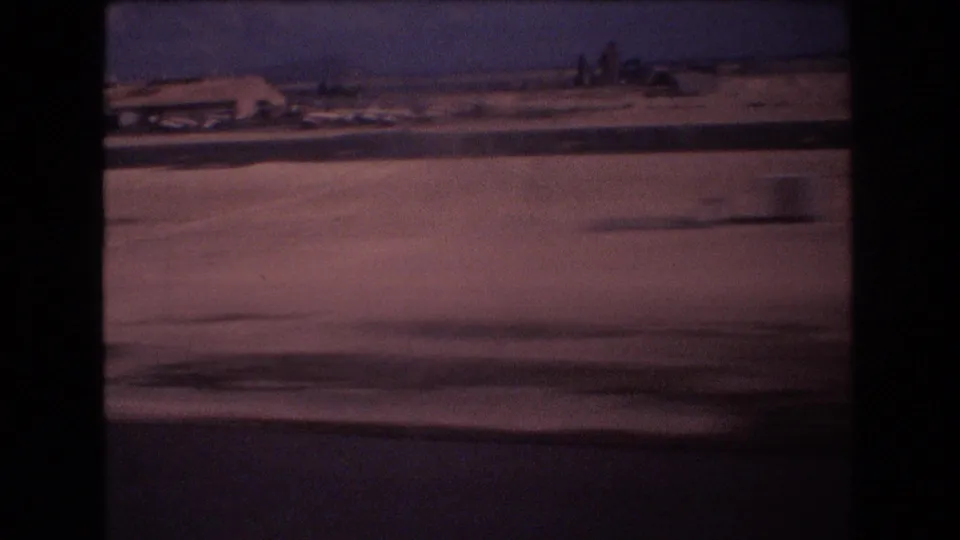 The 1977 home-movie frame appears to show an arid roadside near Honolulu with distant low buildings, sparse trees, and the car-window edge visible in the foreground.