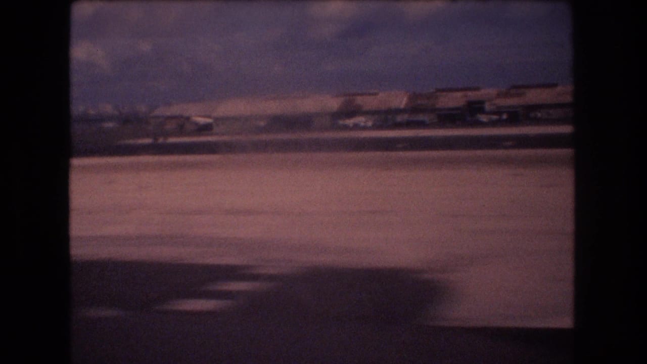 1977 Honolulu home-movie frame showing runway pavement, distant airport structures, and a hazy sky from a moving passenger viewpoint.