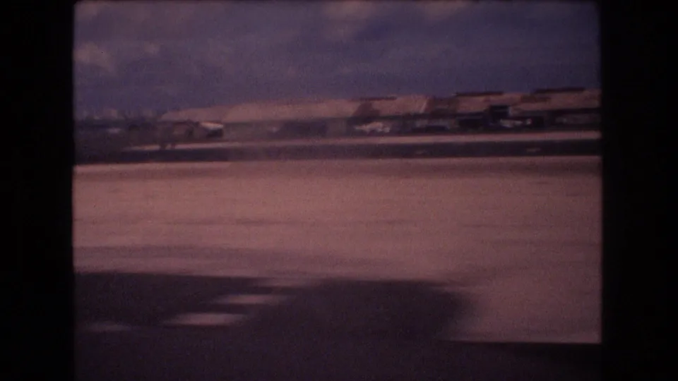 1977 Honolulu home-movie frame showing runway pavement, distant airport structures, and a hazy sky from a moving passenger viewpoint.