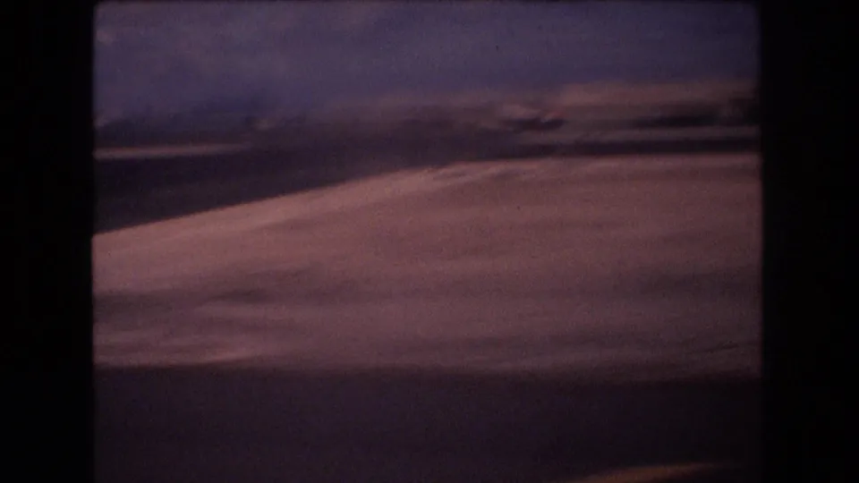 1977 Honolulu airport footage looking across runway pavement with a soft aircraft wing edge, distant airport structures, and hazy sky.