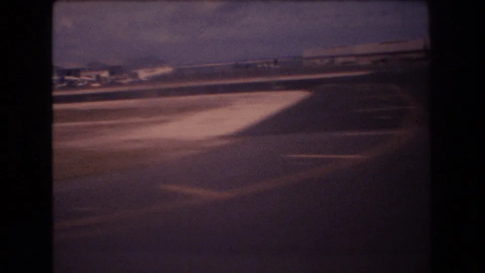 1977 Honolulu home-movie frame with runway or taxiway markings, distant terminal buildings, and a low moving view across the airport surface.