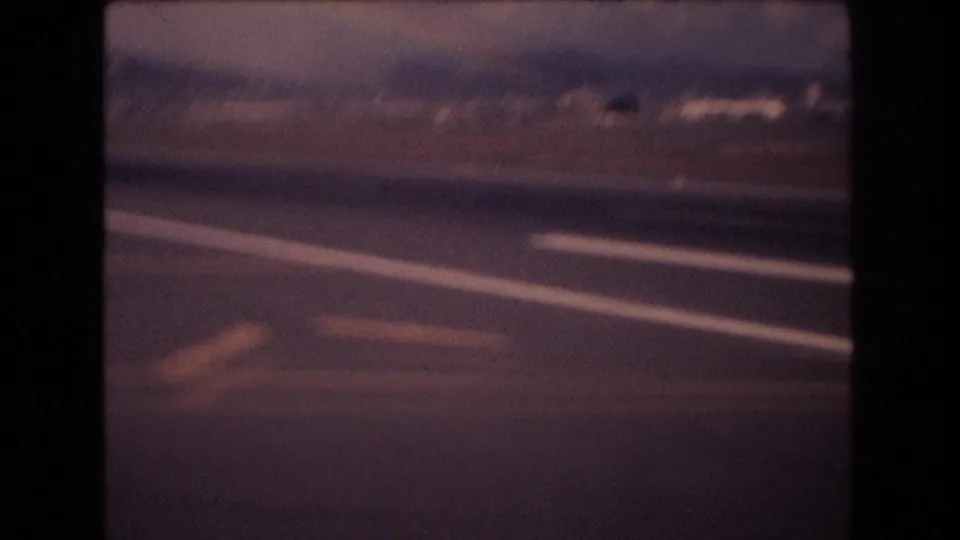 1977 Honolulu airport frame showing blurred runway striping, tarmac pavement, distant structures, and warm 8mm color from a moving travel perspective.