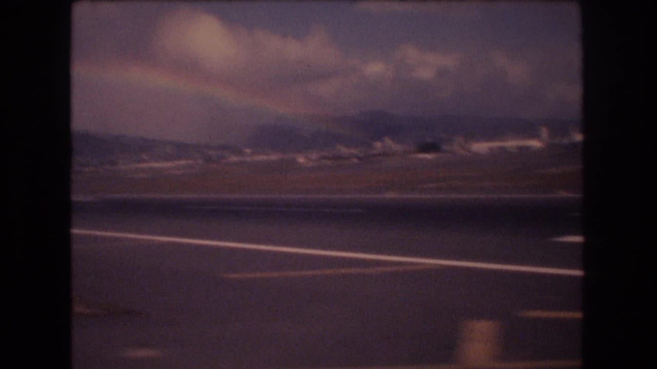 Honolulu airport runway viewed from inside an aircraft in 1977, with distant mountains, heavy clouds, and a faint rainbow-like band across the sky.
