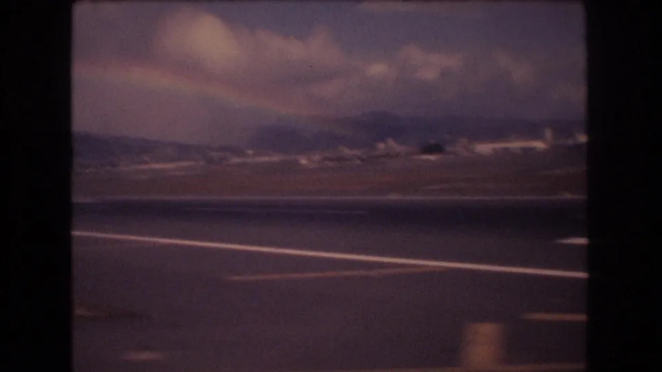 Honolulu airport runway viewed from inside an aircraft in 1977, with distant mountains, heavy clouds, and a faint rainbow-like band across the sky.